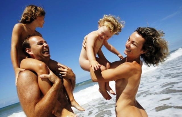 Famille souriante (papa, maman et deux jeunes enfants) sur une plage de sable fin au Pradet, mer turquoise, parasol, ambiance joyeuse.